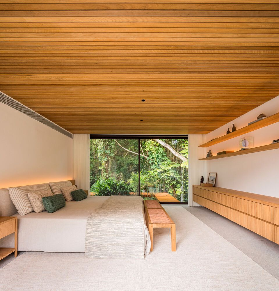 Master bedroom: timber-lined ceiling, white bedding, timber bench at foot, floating timber shelves, floor-to-ceiling glass to forest