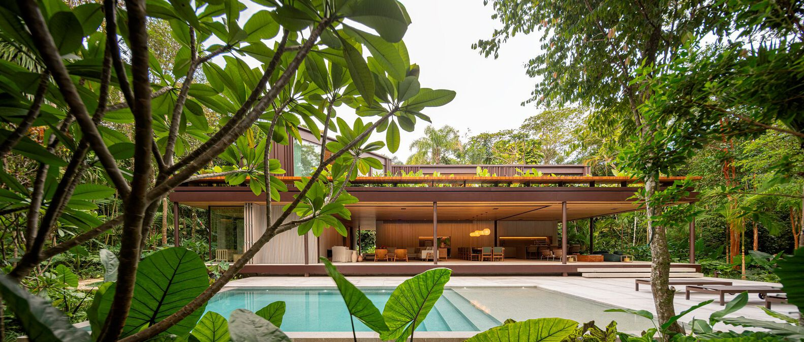 Pool and rear facade: open ground floor under timber ceiling, two upper volumes visible, tropical garden framing the pool