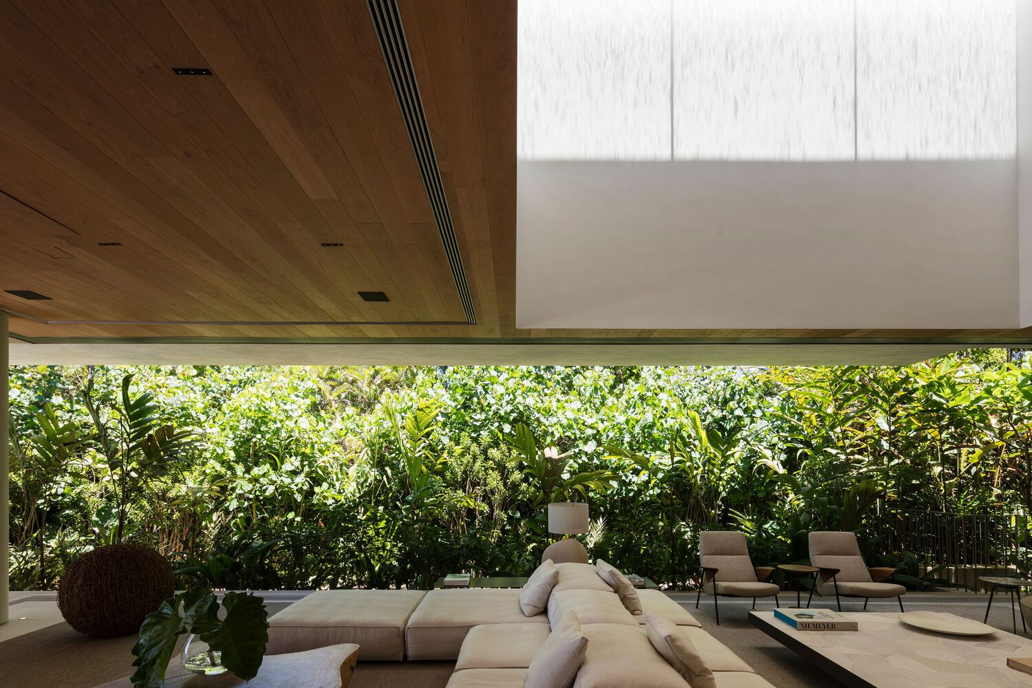 Timber ceiling plane meeting white volume above horizontal glazing overlooking garden and furniture