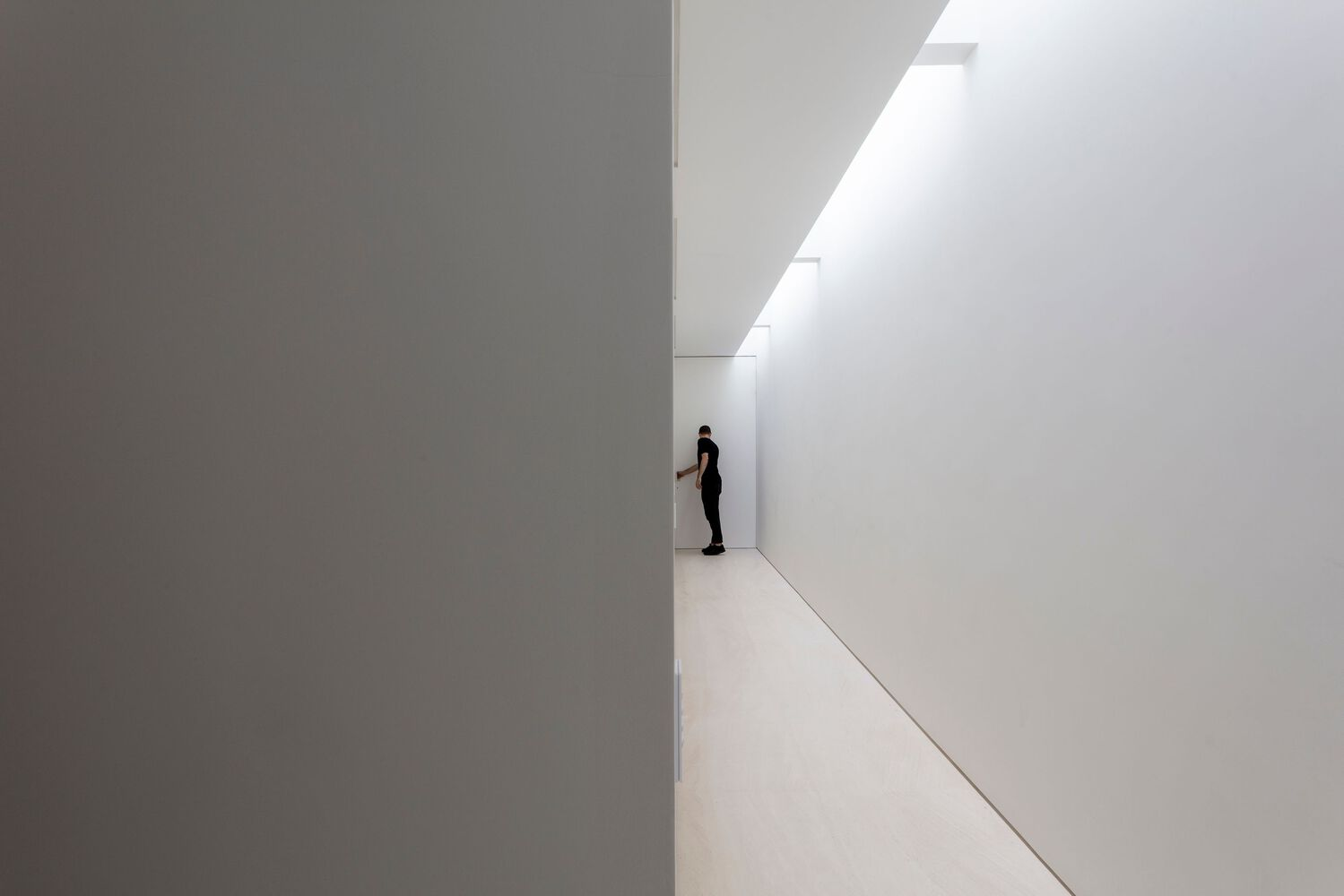 White corridor with a single figure standing at the far end beneath a clerestory window