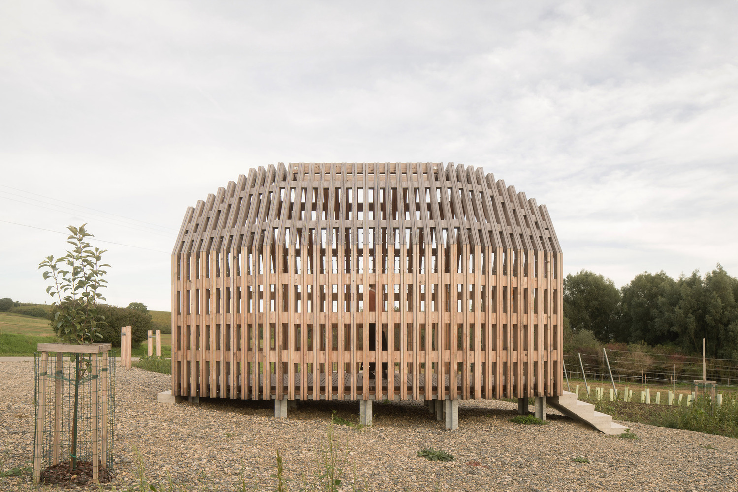 Curved timber slat facade with open gable end facing gravel courtyard and distant tree line