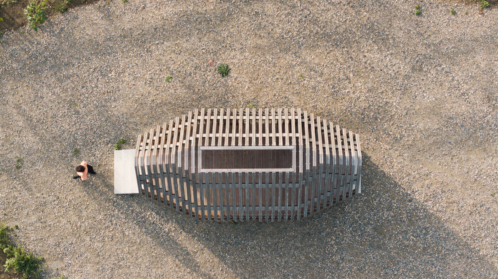 Top-down view of oval timber slat structure with rectangular opening on gravel field