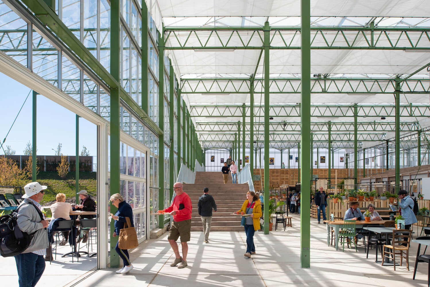 Timber staircase rising through a glass-roofed atrium with green structural beams and seated visitors below