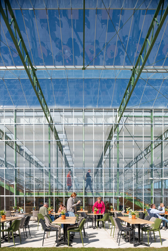 Dining area beneath a transparent roof with green steel structure and visitors at wooden tables in afternoon light
