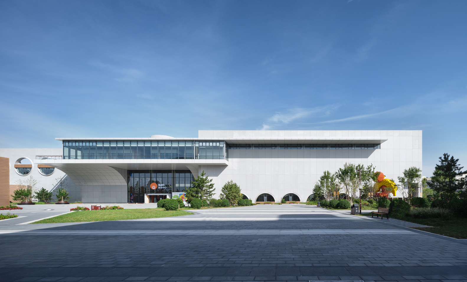 Sports centre approach: white and brick-clad multi-storey volume with cantilevered balconies, landscaped path
