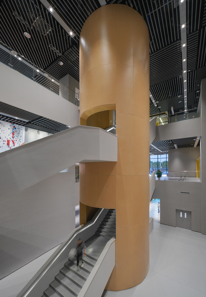Covered walkway: V-shaped concrete columns supporting timber-clad soffit, cultural centre facade with arched and circular openings beyond