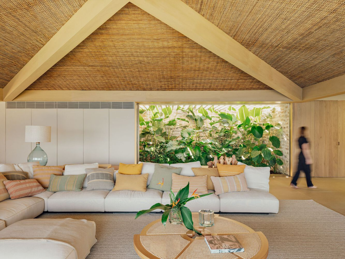 Living room with woven ceiling panels and horizontal window framing planted courtyard with figure passing