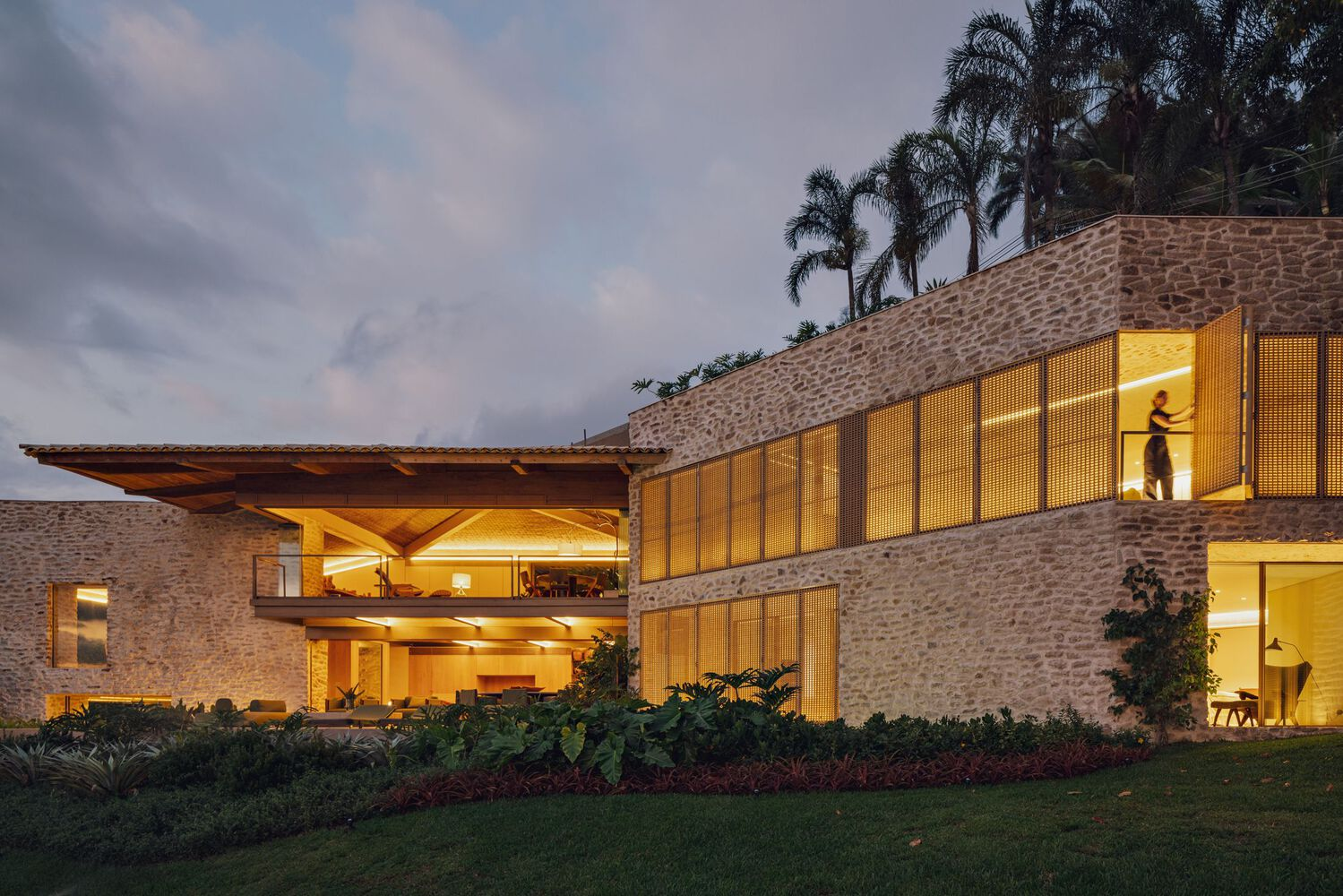 Exterior at dusk showing stone volumes with illuminated timber screens and palm trees on hillside