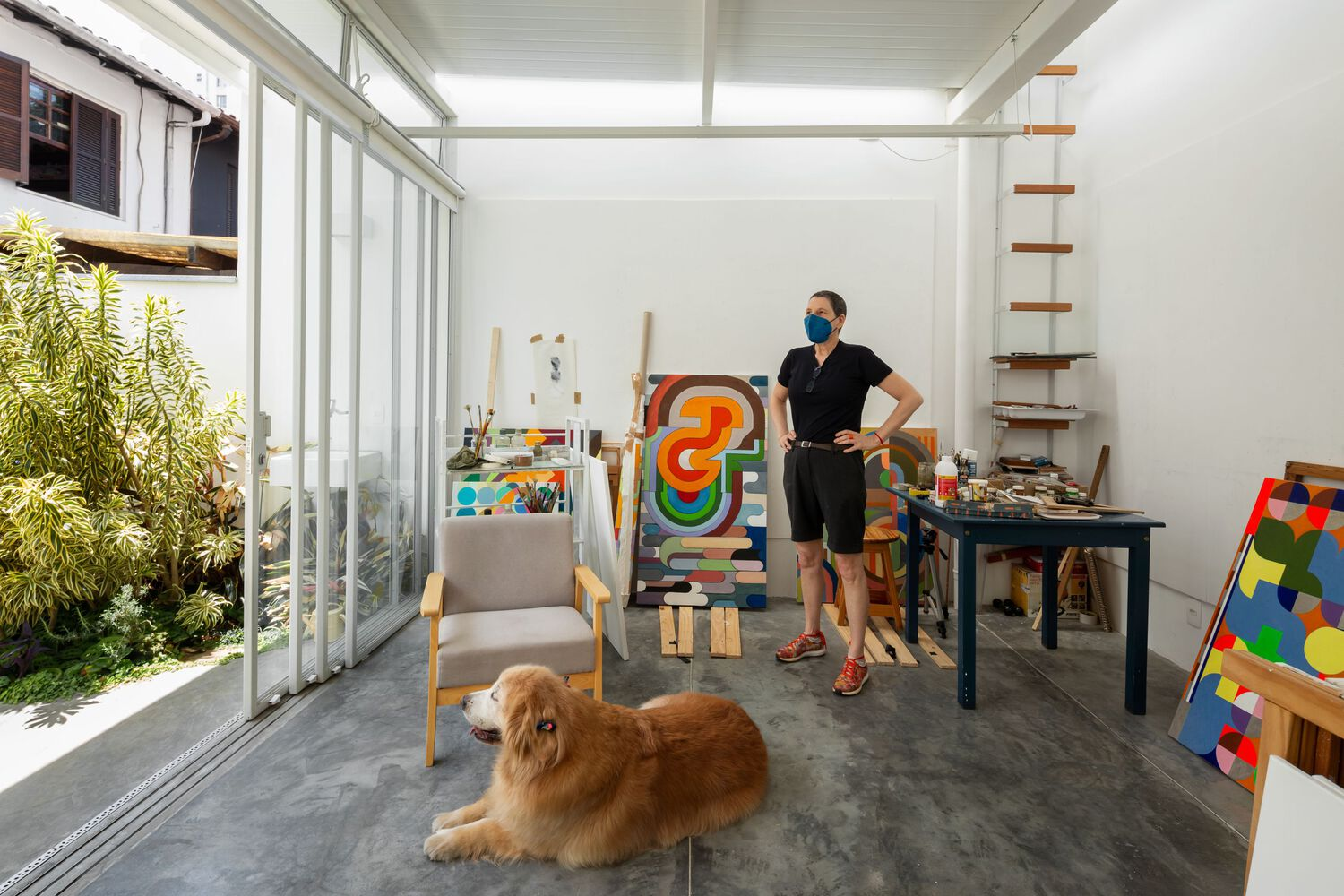 Atelier interior looking out: full-width sliding glass to the garden courtyard, armchairs, easels, paintings