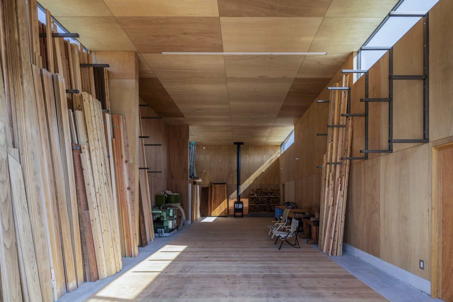 Double-height workshop interior with plywood ceiling panels and stacked timber leaning against the left wall