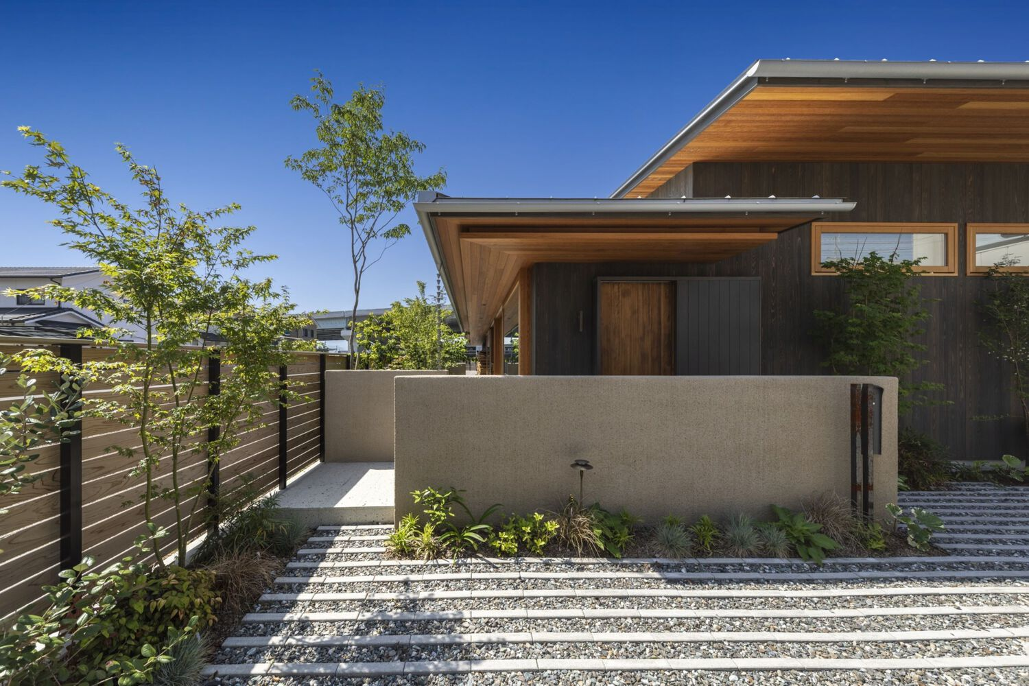 Front approach with concrete wall, striped gravel pathway and young trees under a clear sky