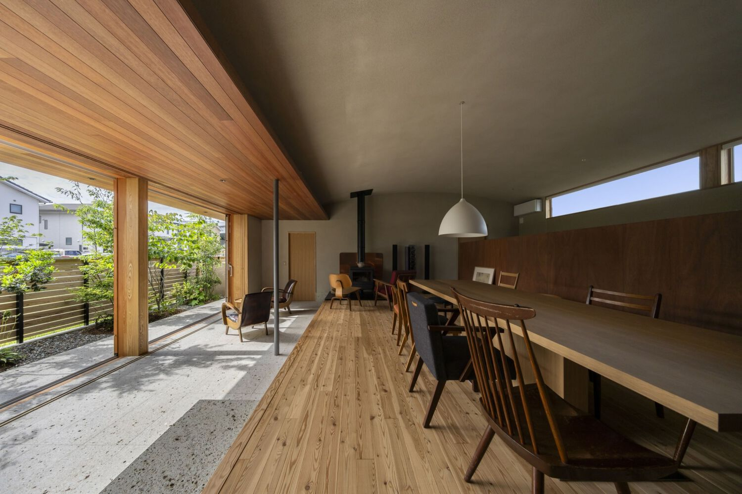 Open-plan interior with timber-clad ceiling, concrete floor, and dining table overlooking a planted courtyard through glazed walls