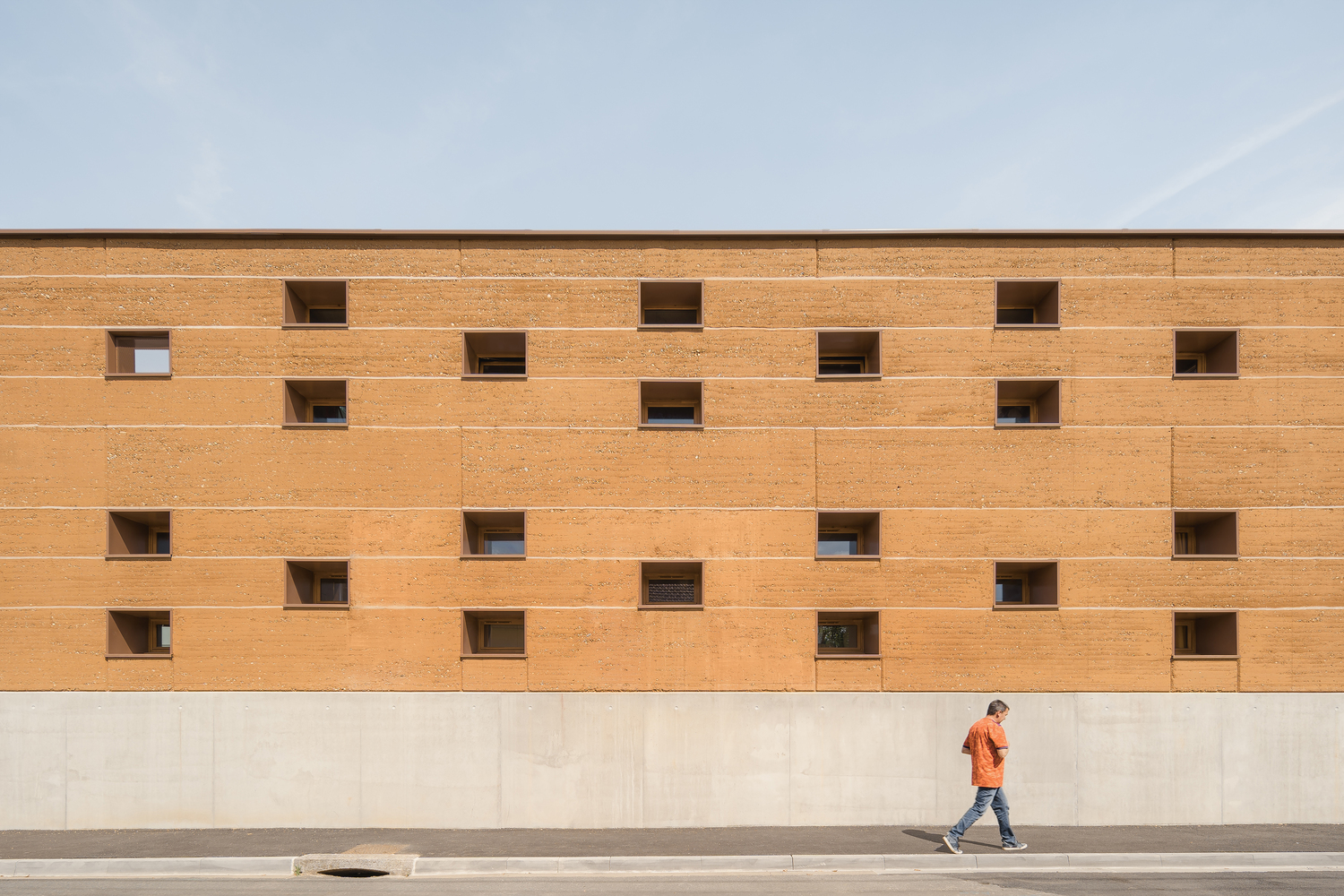 Street facade: long rammed earth wall with scattered small square windows, figure walking past