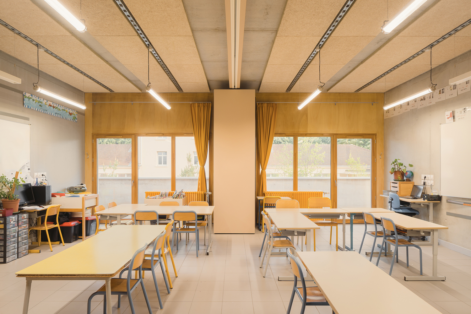 Classroom: plywood wall panels, timber-framed windows with yellow curtains, CLT timber ceiling panels, student desks and coloured chairs