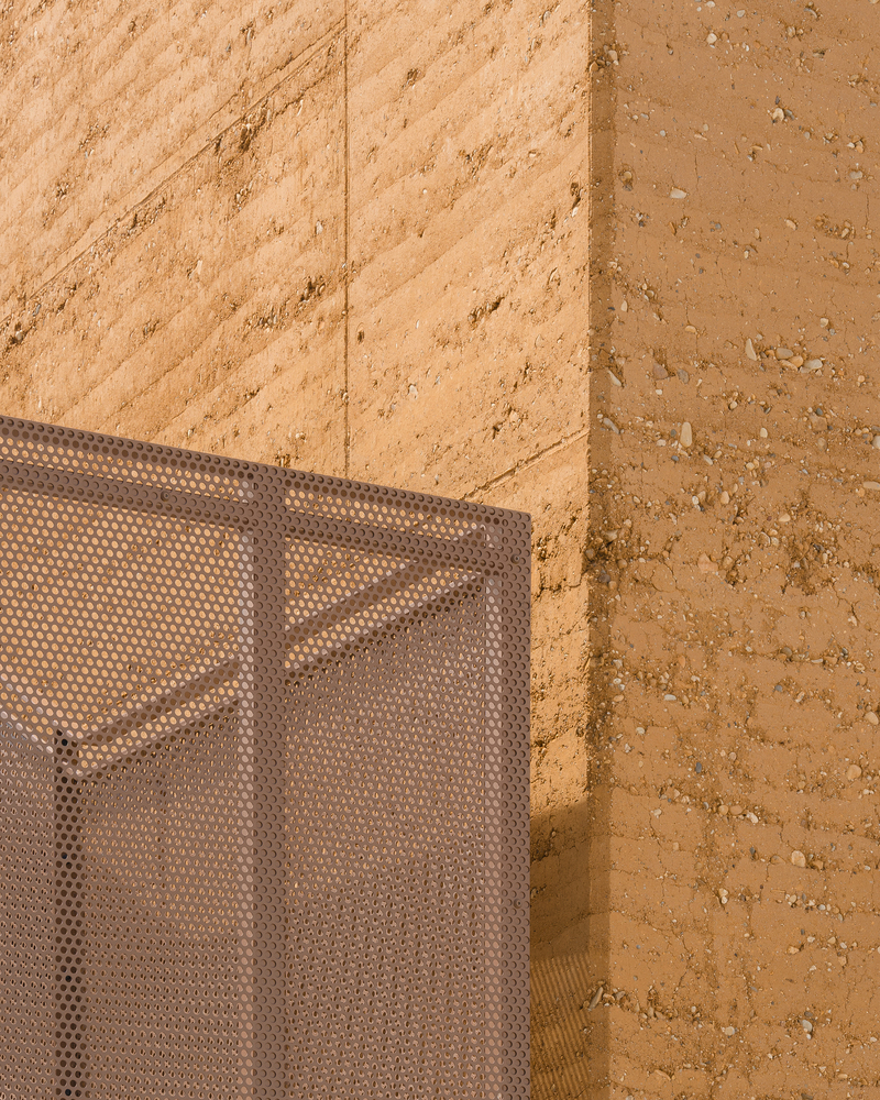 Detail: rammed earth wall corner meeting perforated metal mesh screen, warm afternoon sunlight on the earth texture
