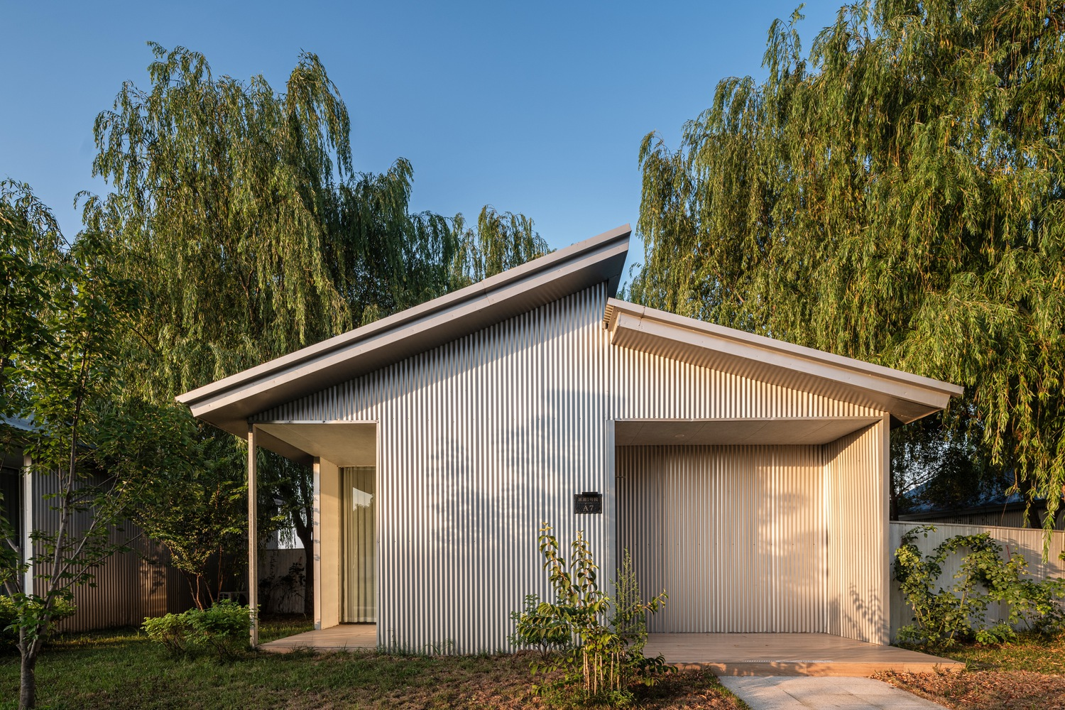 Corrugated metal facade with pitched roof and garage door framed by weeping willow branches