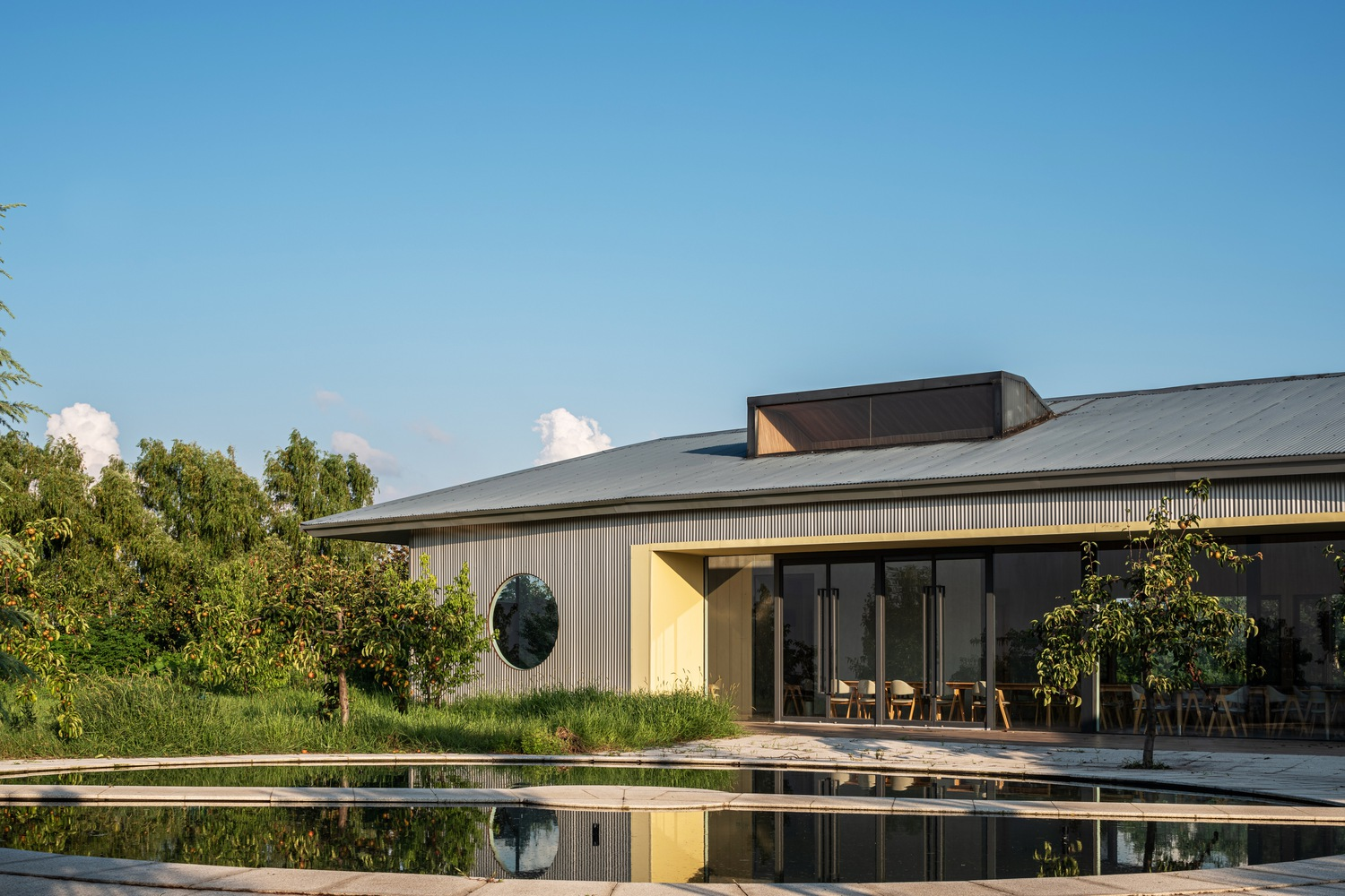 Poolside view of the corrugated metal pavilion with circular window reflected in still water