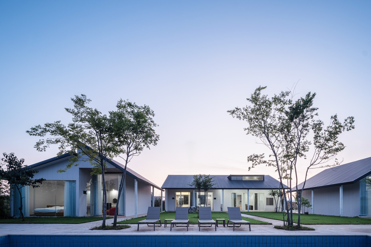 Courtyard with pool and lounge chairs between gabled pavilions framed by young trees at dusk