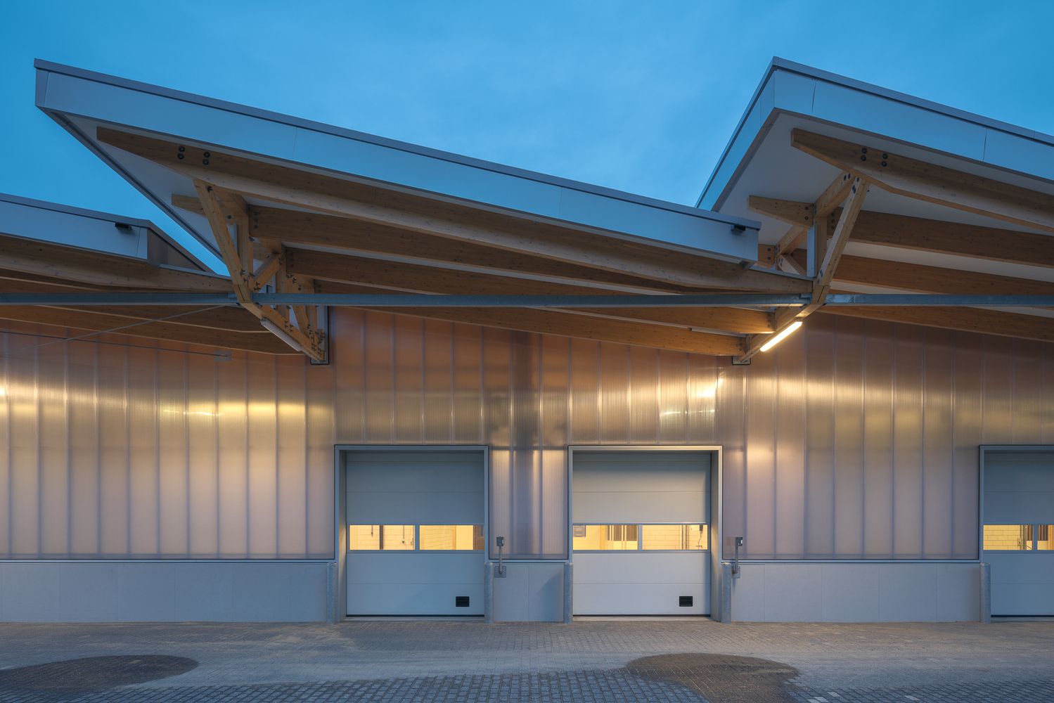 Expedition facade at dusk: timber trusses and canopy lit from below, three roller doors closed