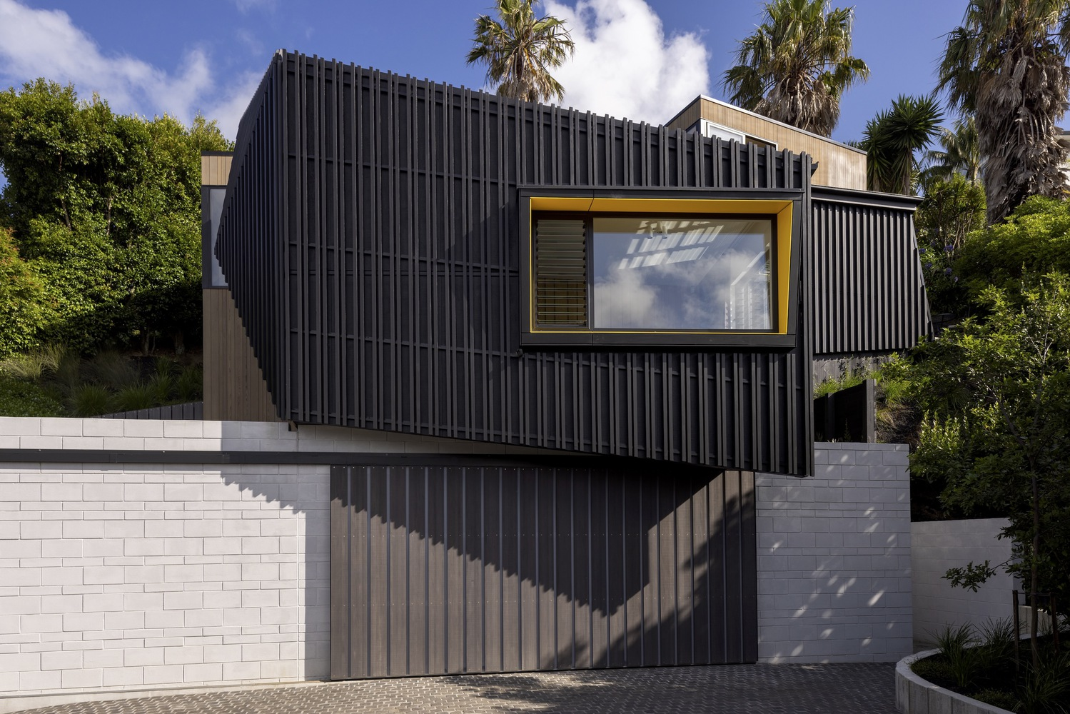 Vertical corrugated metal facade with yellow-framed window above white block retaining wall and palm trees