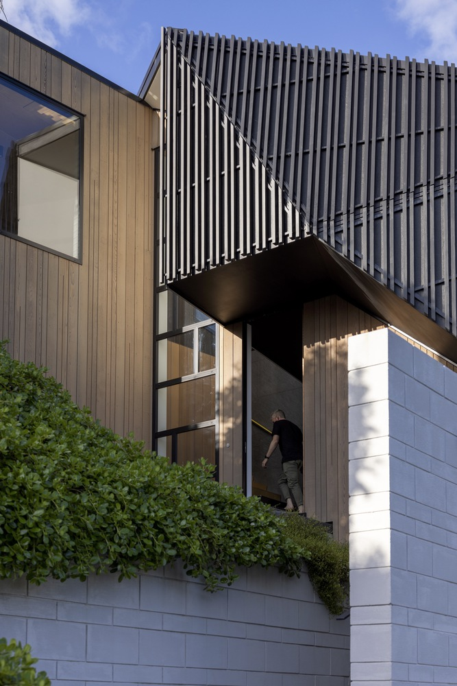 Cantilevered upper volume with vertical metal screening above timber-clad bridge and white tiled base