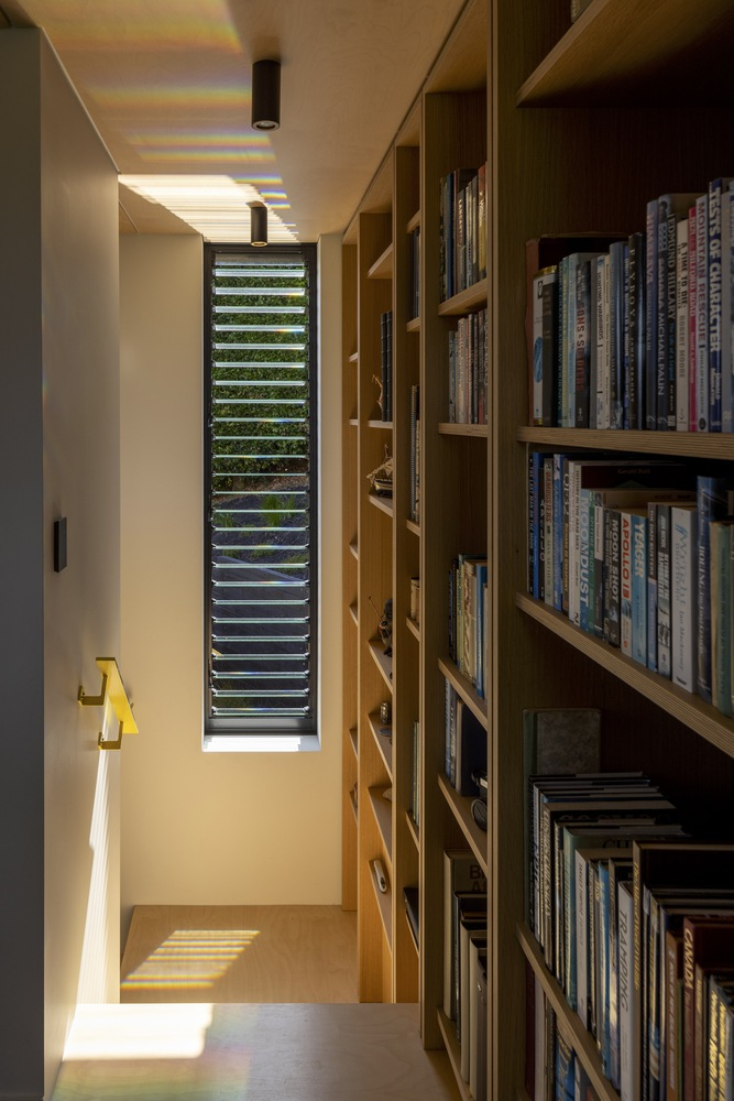 Narrow corridor with timber bookshelves and a vertical louvered window casting striped sunlight across the floor