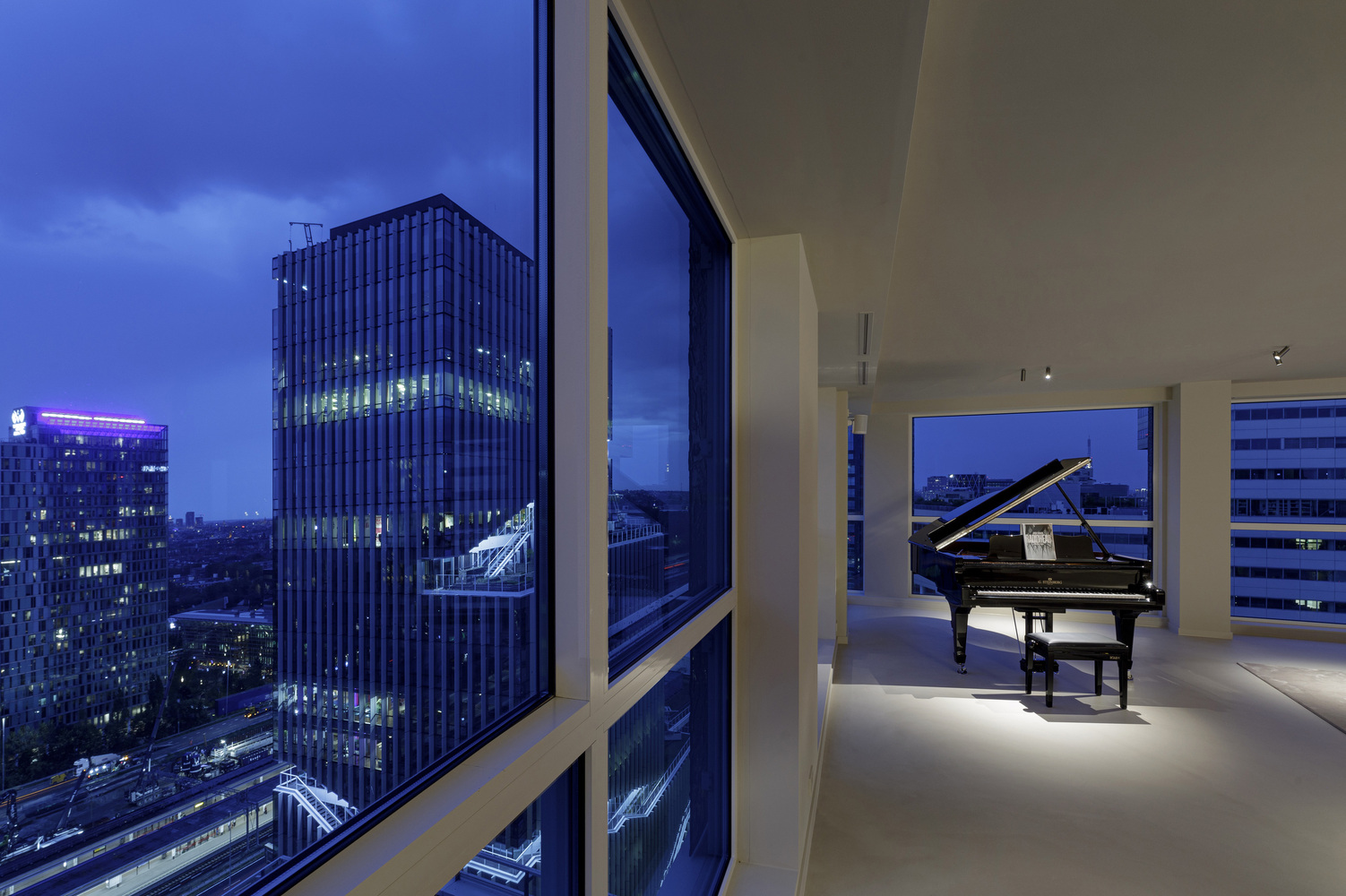 Floor-to-ceiling windows framing the city at dusk with a grand piano illuminated by floor uplights