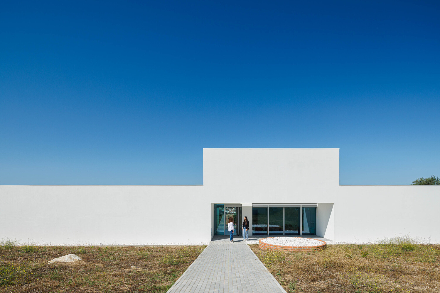 White stucco facade with recessed glazed entrance and paved walkway through dry grasses under clear sky