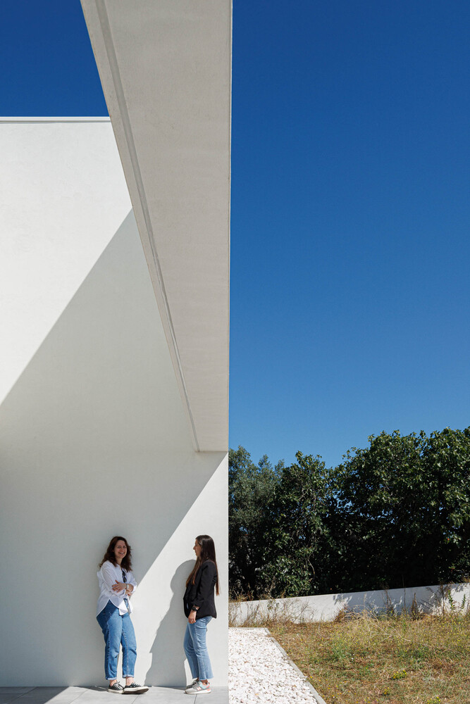 Two figures standing beneath a cantilevered white overhang with geometric shadows on a sunny day