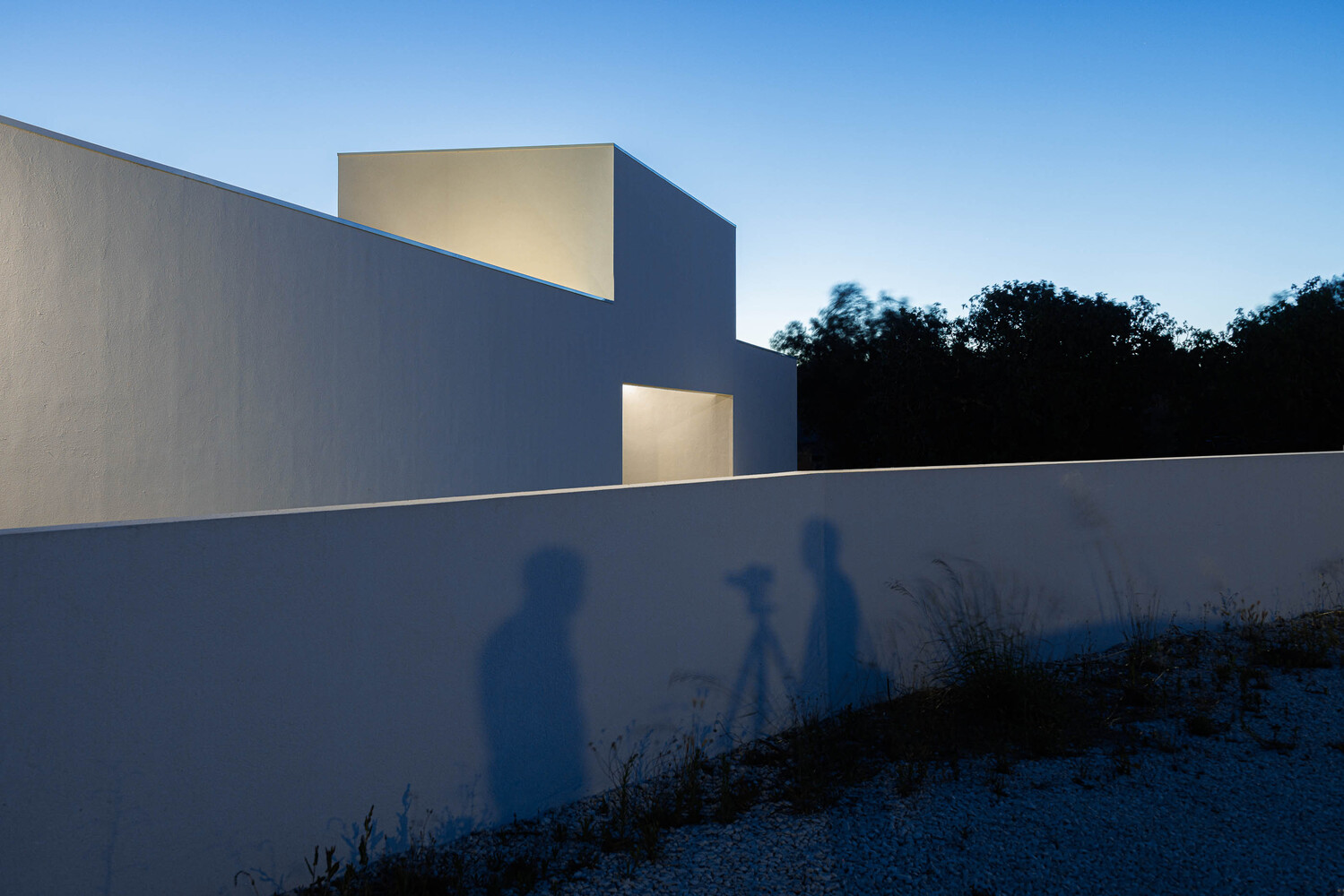 White geometric volumes with perforated openings casting shadows of visitors on a gravel path at dusk