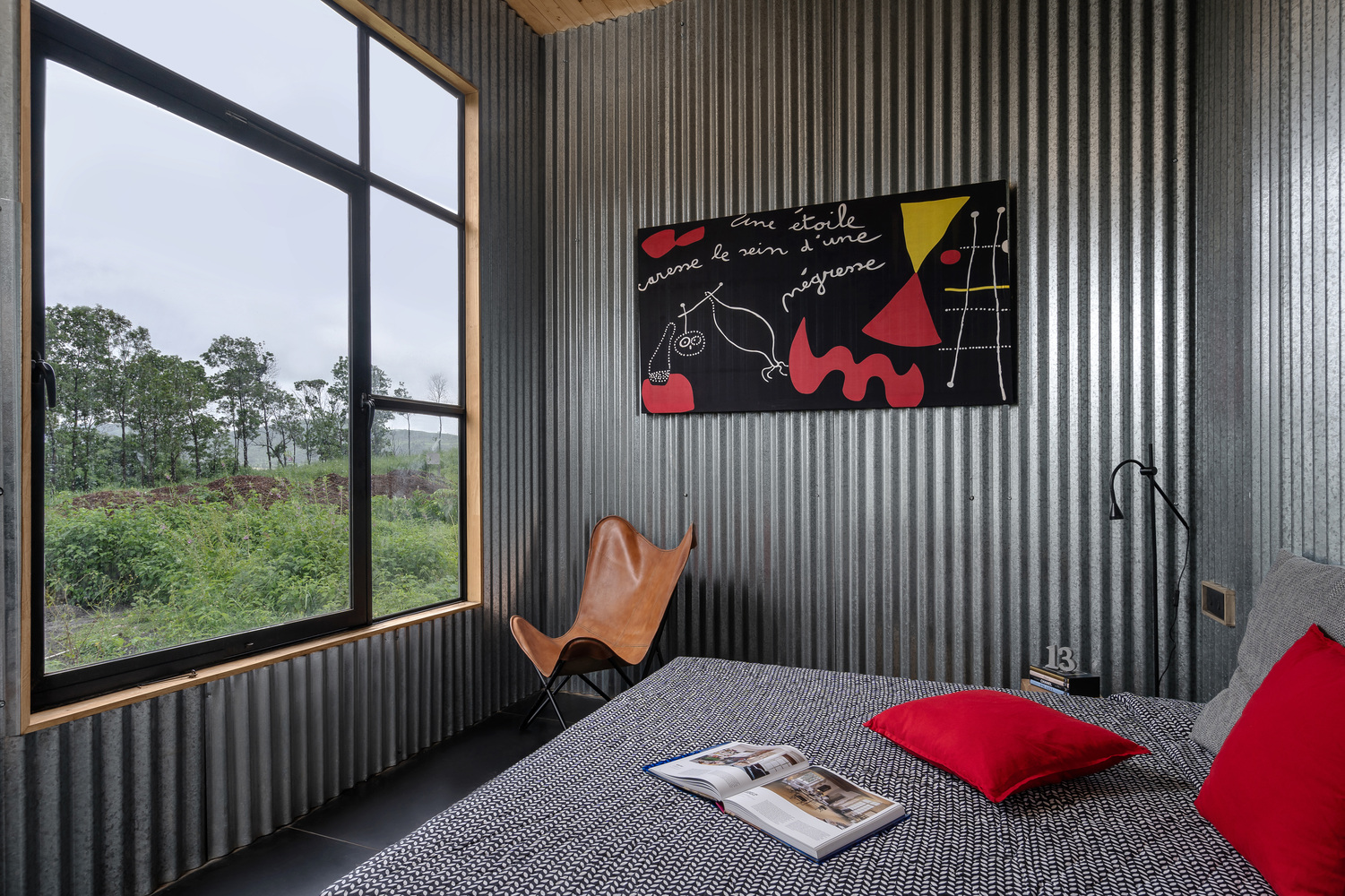 Bedroom with corrugated metal walls, black-framed window, and leather butterfly chair beside the bed
