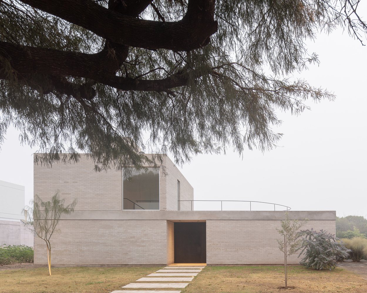 Garden approach: the two-storey brick volume framed by a mature tree, overcast sky