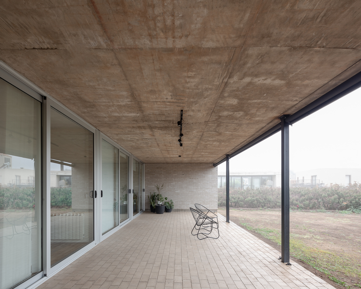 Covered porch: exposed concrete ceiling, brick floor, wire chair, full-height glass to garden