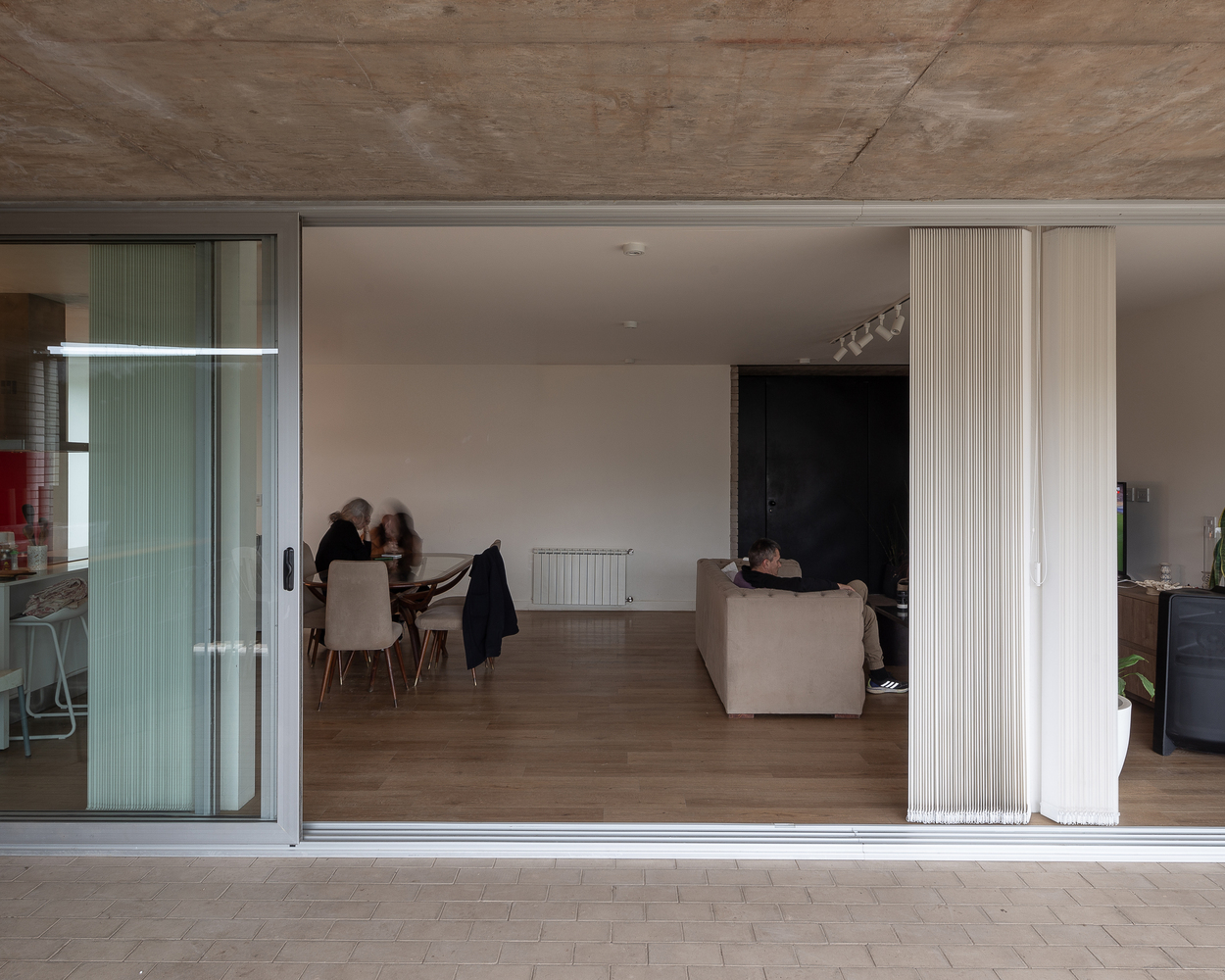 Interior through glass: open-plan living and dining, family at table, concrete ceiling