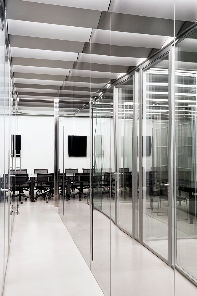 Glass partition corridor: stainless steel frames, meeting room visible beyond, black chairs, monochrome palette
