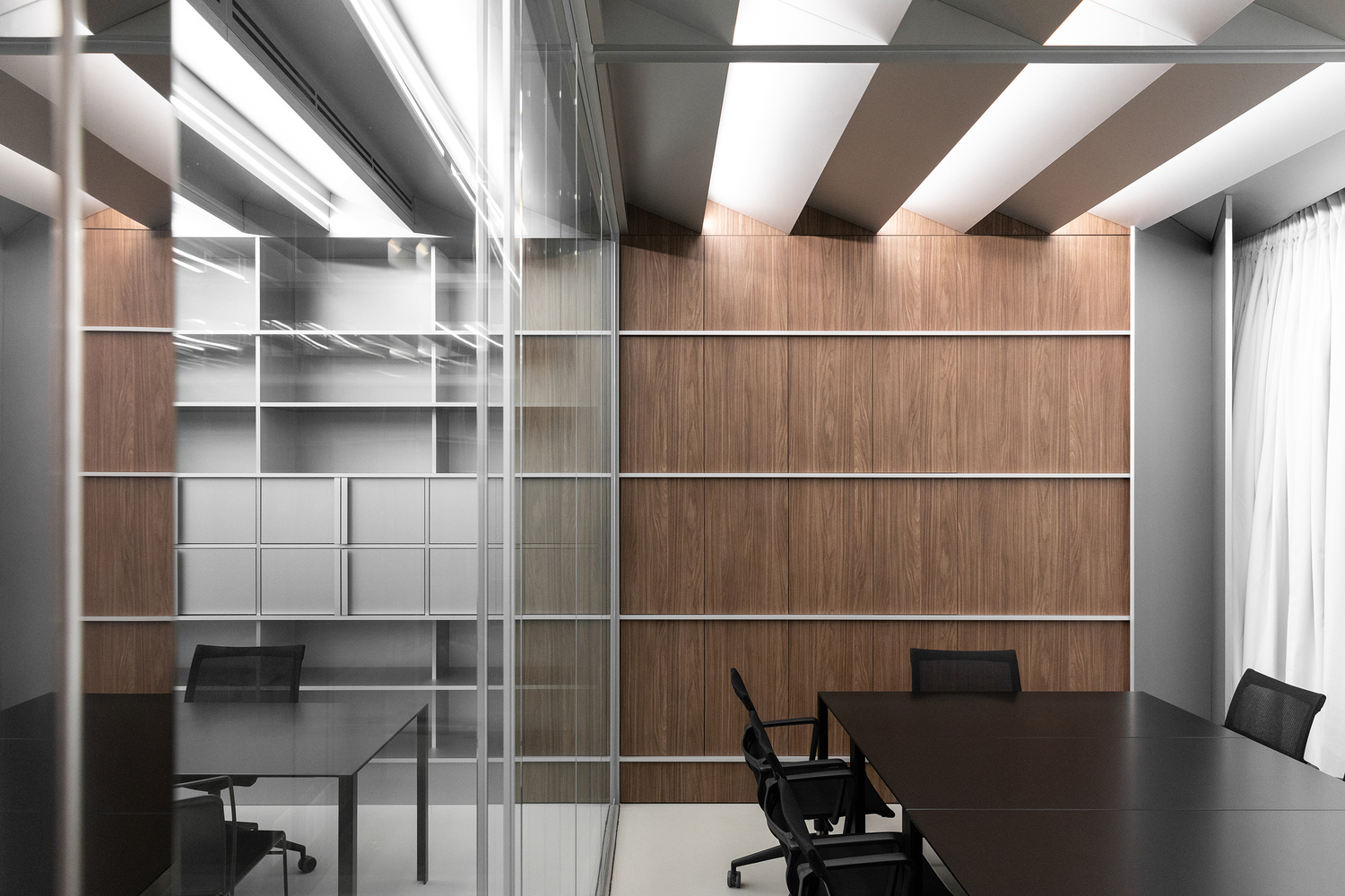 Private office through glass: walnut wall, grey open shelving unit, black desk, ceiling baffles above