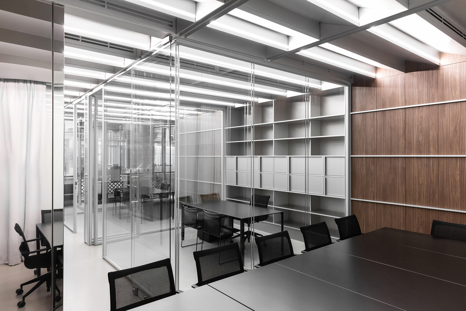 Open office: glass partitions layered in depth, grey shelving, walnut wall, black table and chairs, curtains at windows