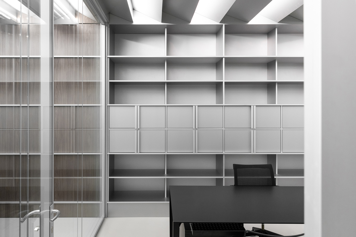 Private office: grey open shelving wall unit filling the back wall, black desk and chair, glass partition at left