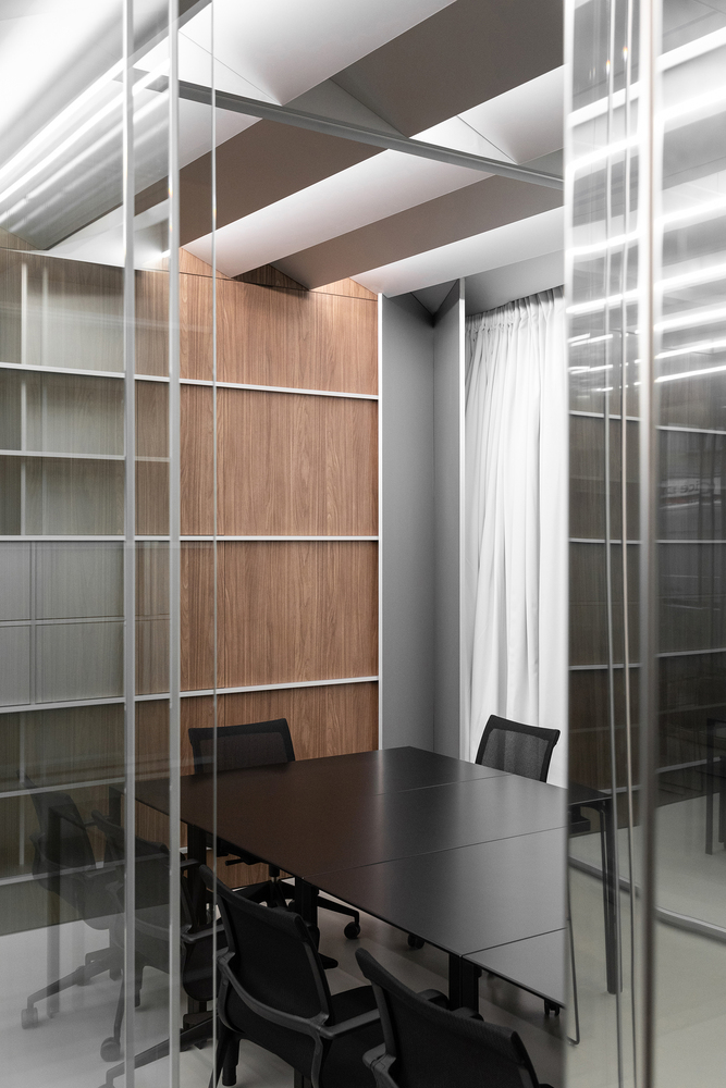 Through glass partition: walnut wall, grey shelving, dark meeting table, sheer curtain at the window