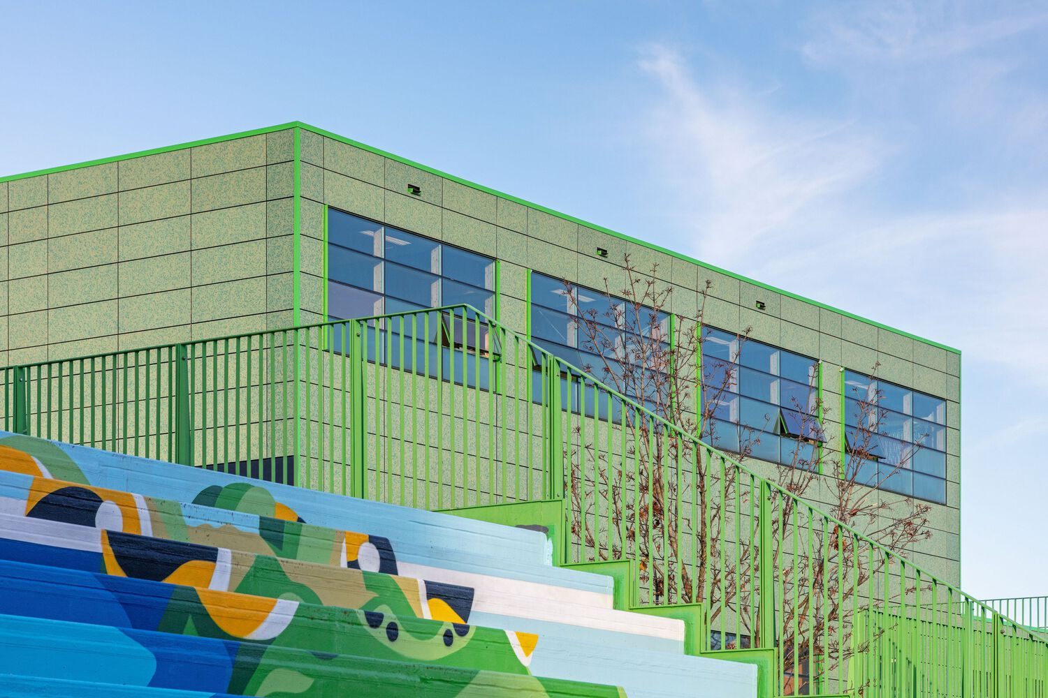 Detail of green metal railing and paneled facade above painted mural with young planted trees