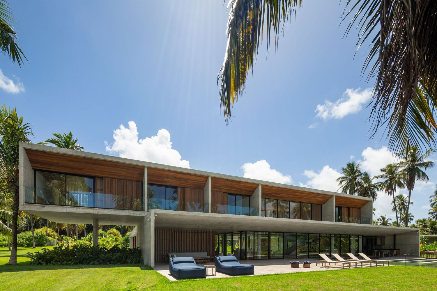 Garden facade: upper bedrooms with deep concrete balconies, timber screens, blue loungers on the lawn