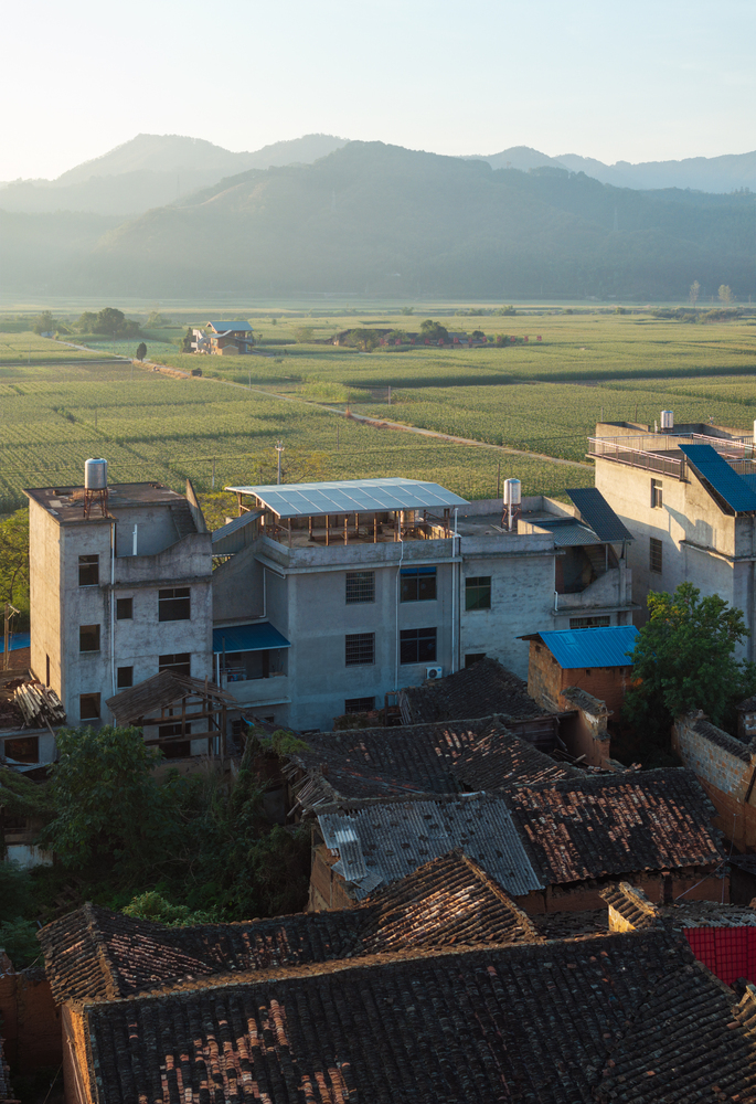 Aerial view of concrete and brick village rooftops with mountains and agricultural fields in morning mist