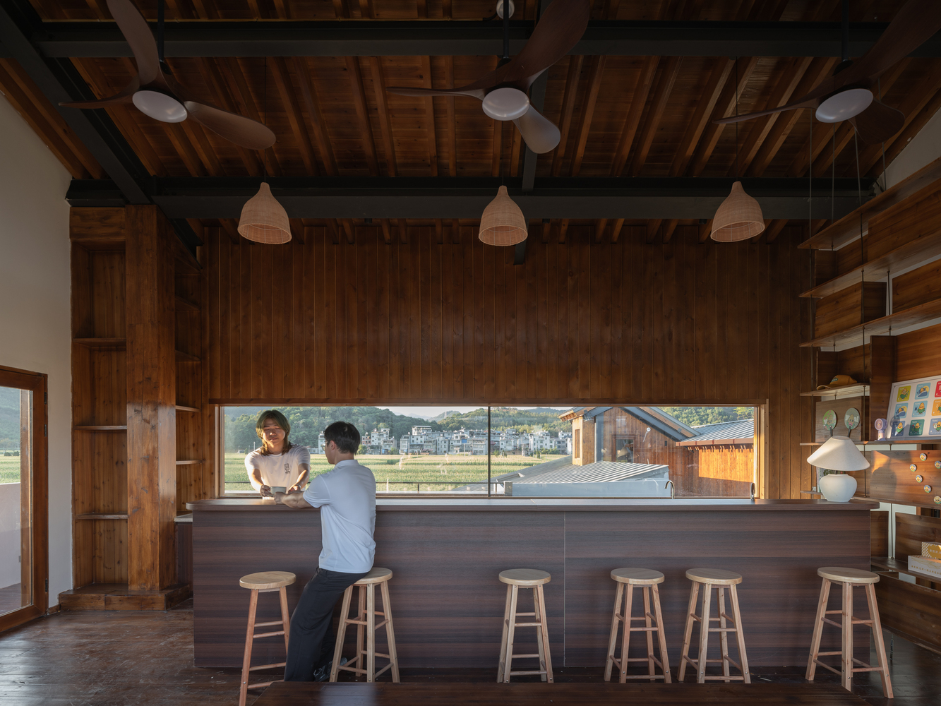 Kitchen counter with timber bar stools and horizontal window framing farmland beyond where two figures stand