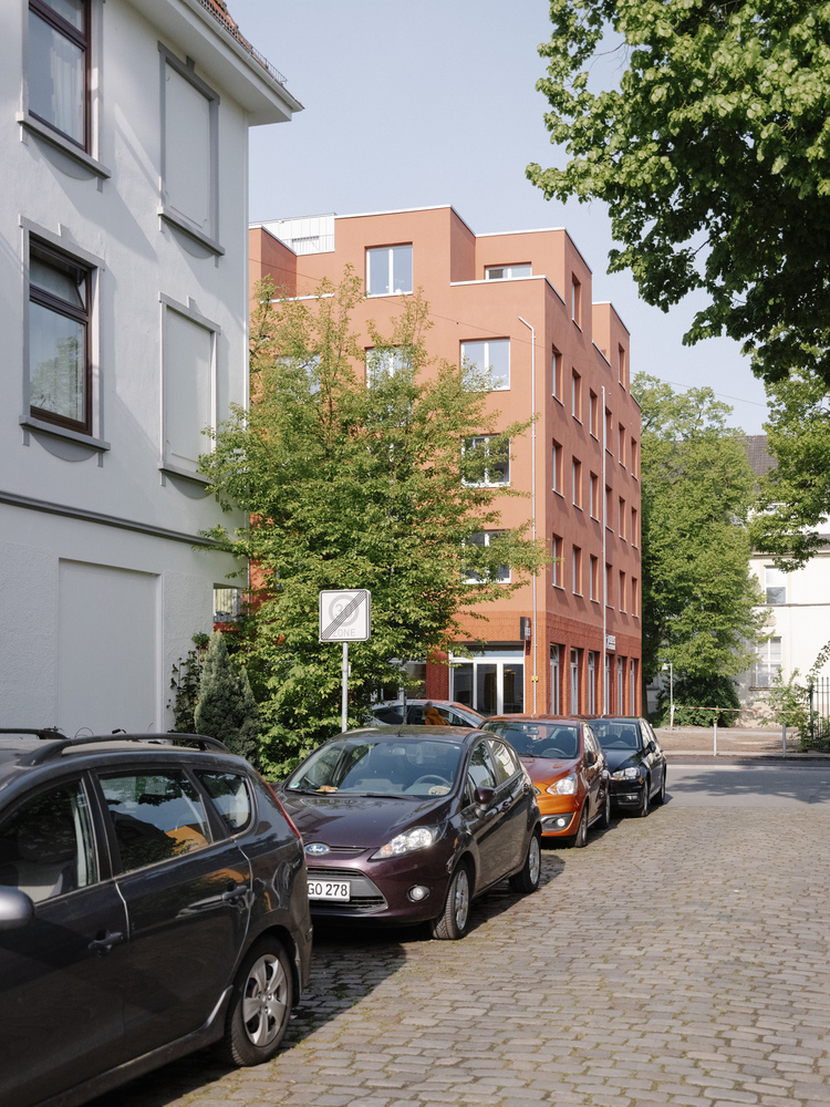 Cobblestone street view with parked cars, mature trees, and terracotta housing block beyond white neighboring building