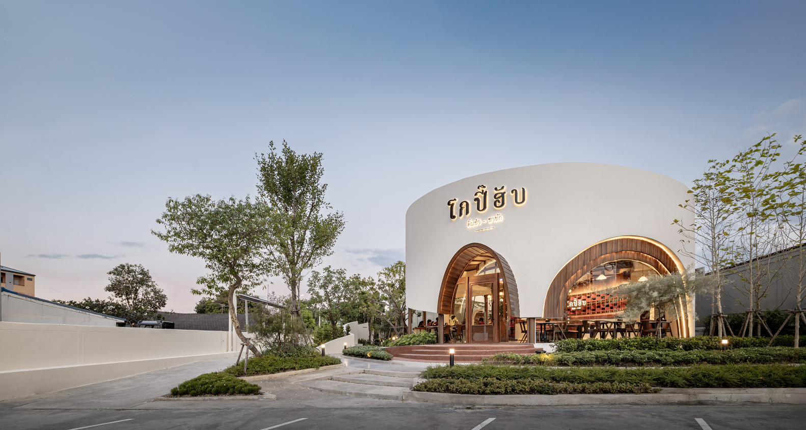 White curved facade with timber-clad arched openings and signage at dusk surrounded by young trees