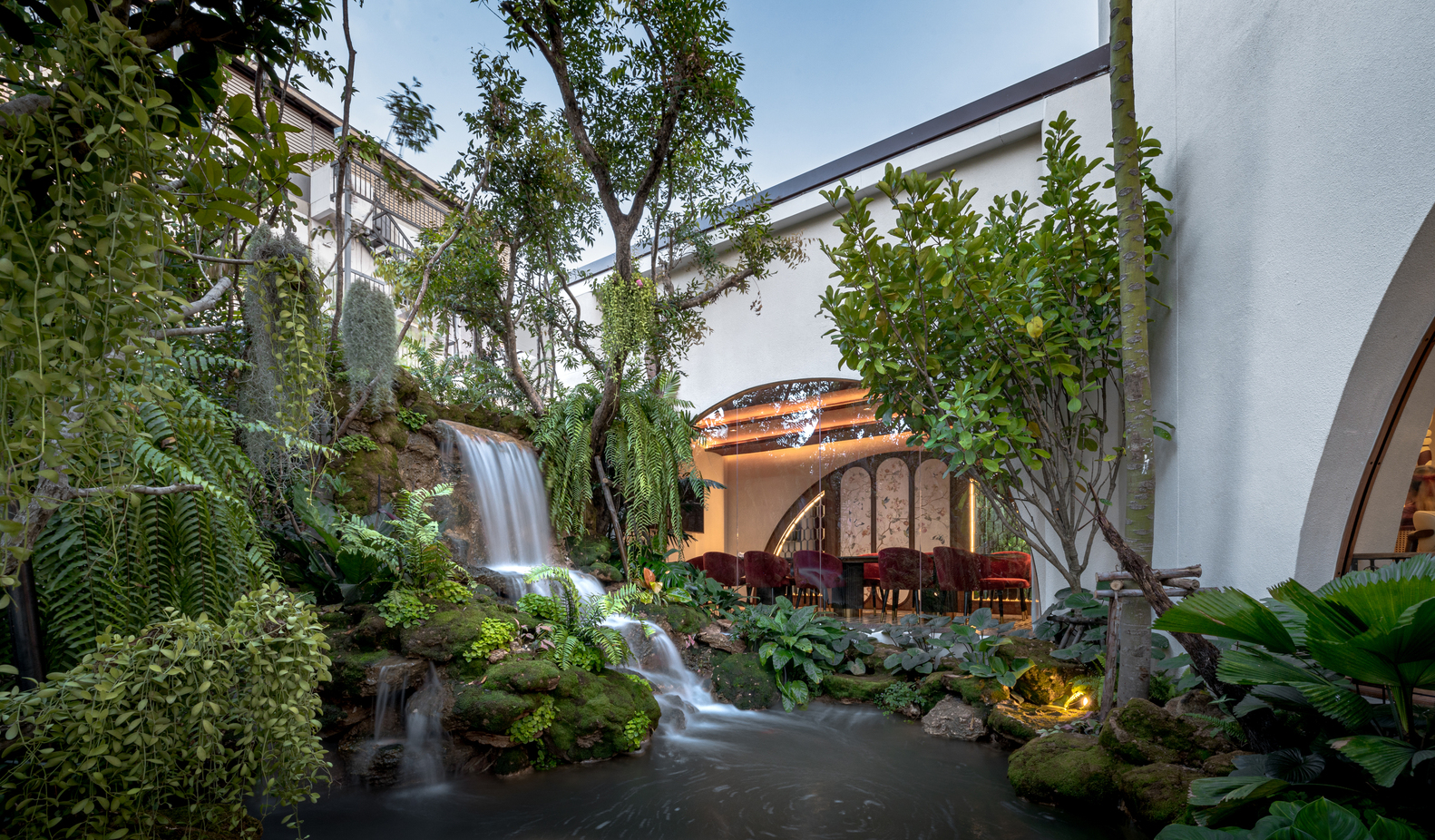 View across pond toward dining pavilion with waterfall and planted landscape at dusk