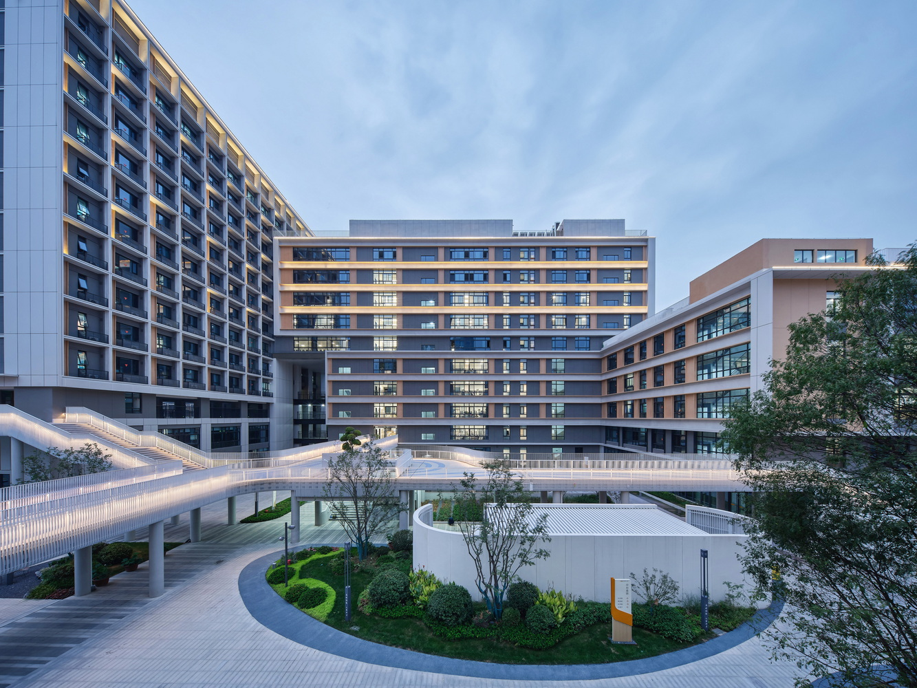 Residential courtyard with curved landscaped beds and an illuminated pedestrian bridge at twilight