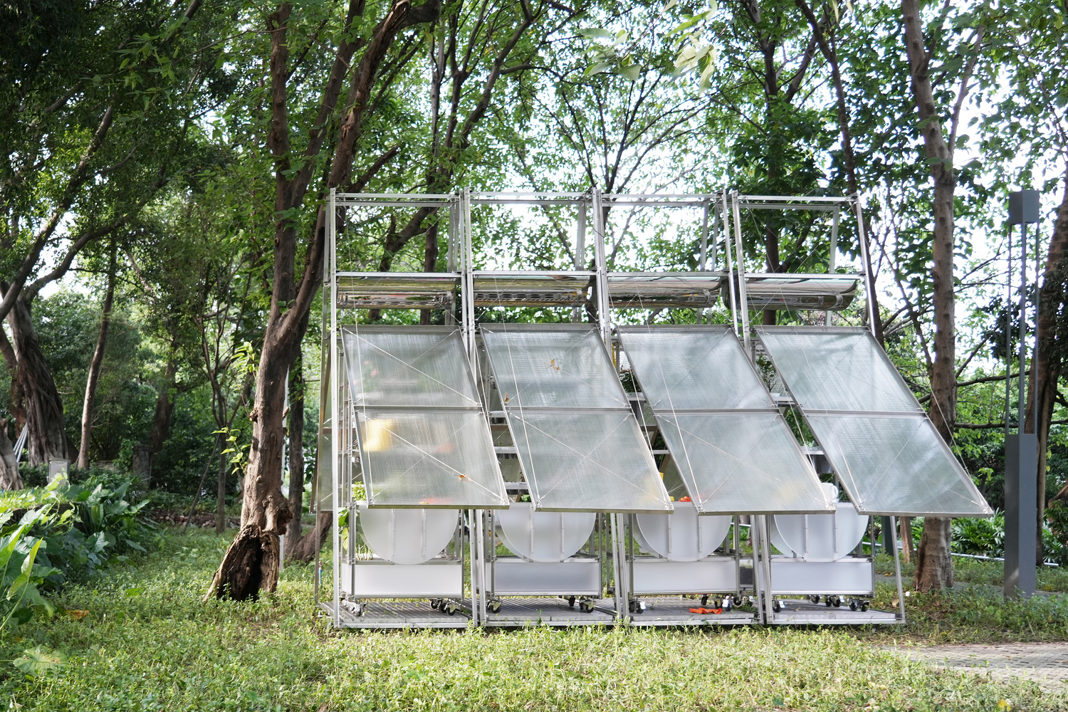 Four angled translucent polycarbonate panels in a white steel frame surrounded by dense green trees