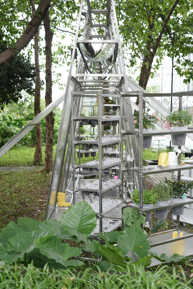 Pyramidal aluminum frame greenhouse structure with tiered planting trays nestled among trees in a park