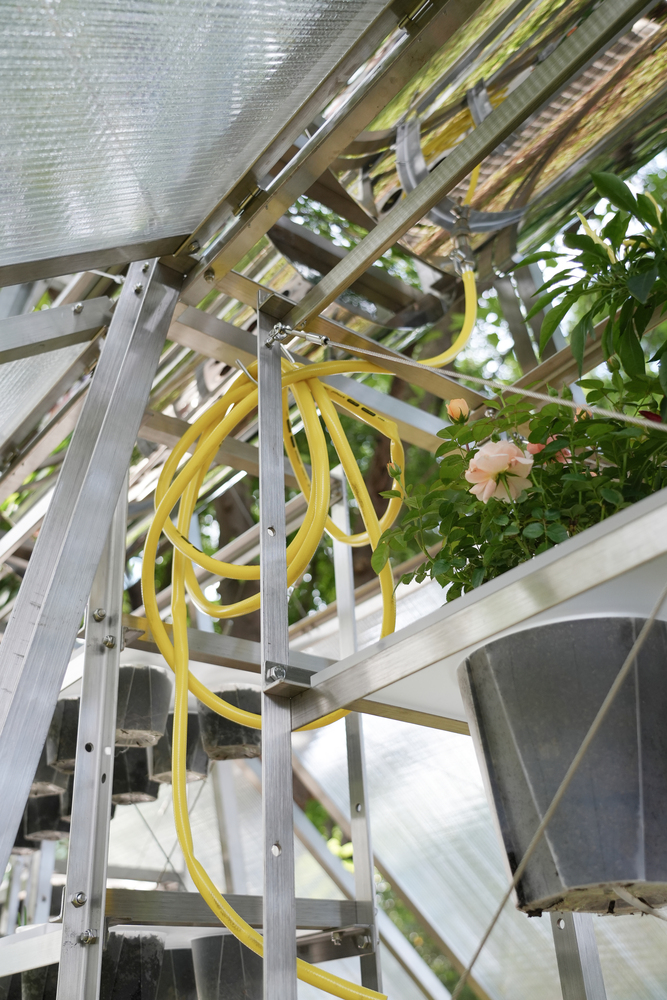 Interior corner showing yellow irrigation tubing coiled around aluminum framing with planted trays and flowering plants